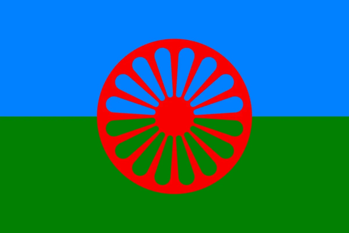 Flagge der Rom*nja und Sinti*zze. Sie ist Blau in der oberen Hälfte und Grün in der unteren Hälfte. Außerdem enthält die Flagge mittig ein Speichenrad. im Zentrum der Flagge.