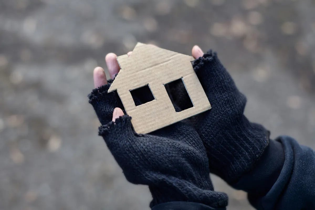Die Hände einer vermutlich obdachlose Person hält mit kaputten, schwarzen Halbhandschuhen ein flaches Haus aus pappe in der Hand.